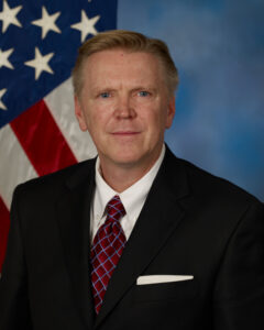Headshot of a man in front of a U.S. Flag. He has short light brown hair. He is wearing a black suit with a white shirt underneath and a red tie.