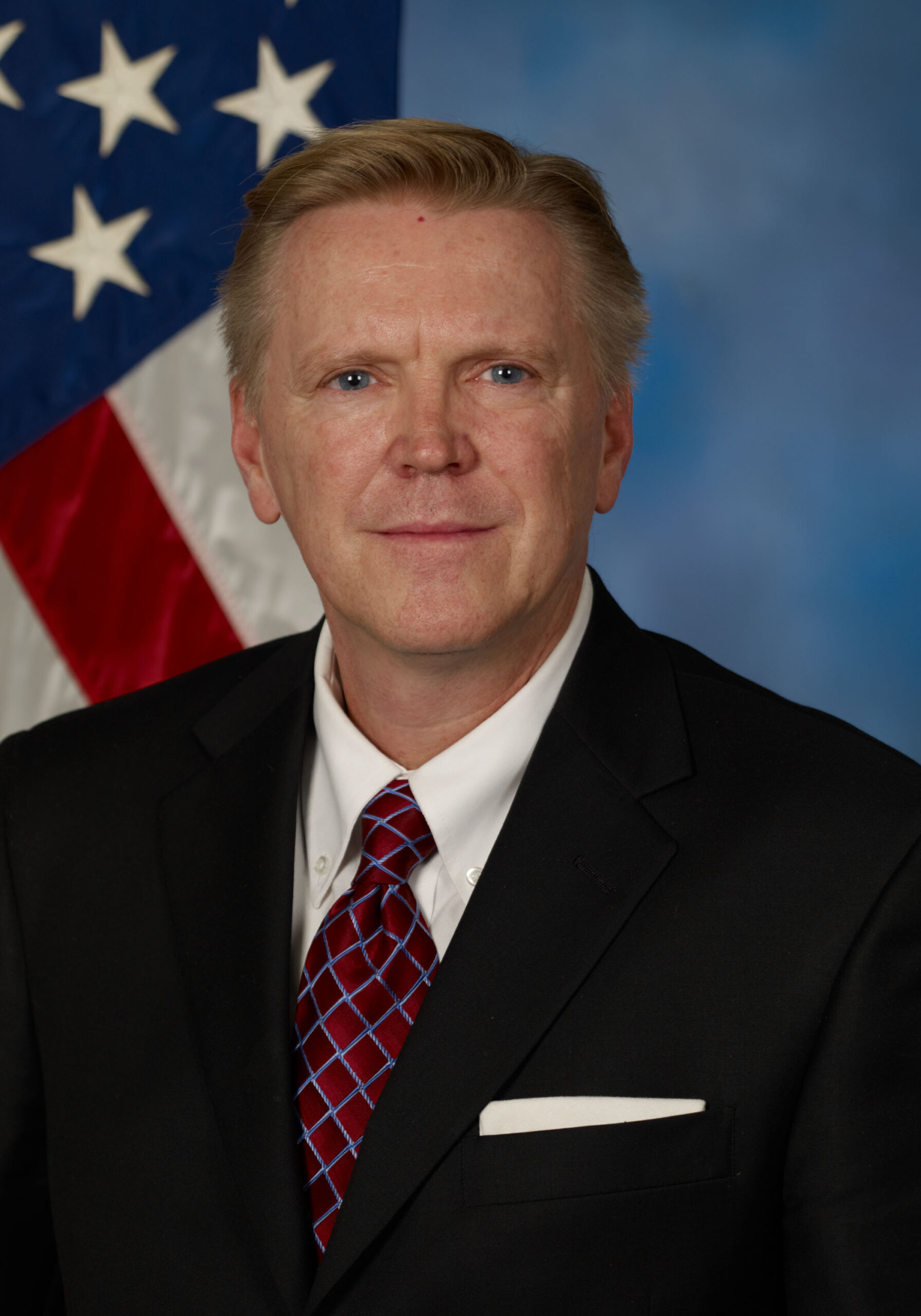 Headshot of a man in front of a U.S. Flag. He has short light brown hair. He is wearing a black suit with a white shirt underneath and a red tie.